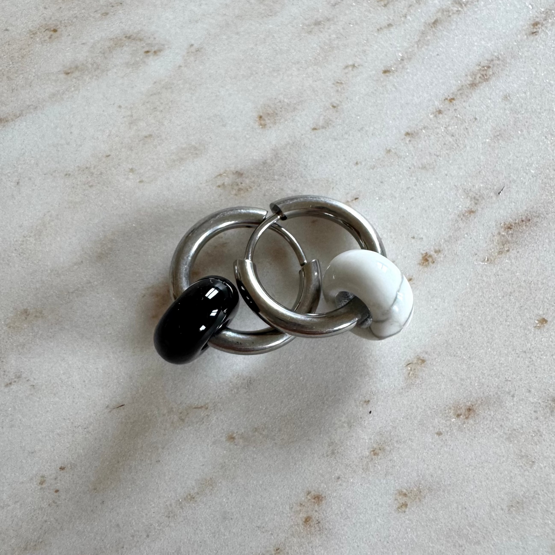 Two silvery hoop earrings on top of each other with 1 black and 1 white donut pendants on beige marble